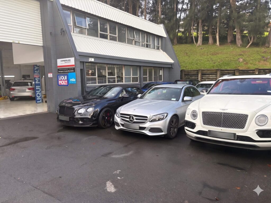 Three luxury cars parked outside an automotive workshop with Liqui Moly branding and a treed background.