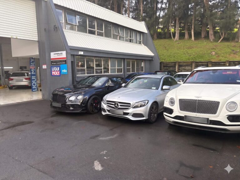 Three luxury cars parked outside an automotive workshop with Liqui Moly branding and a treed background.