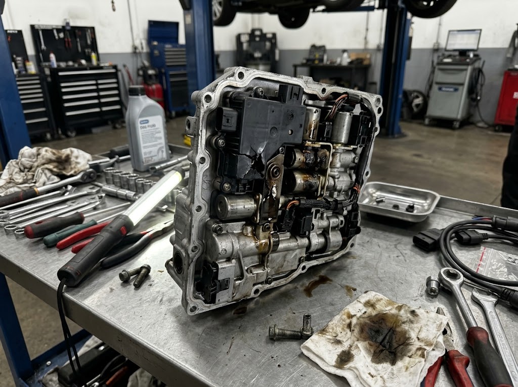 A damaged DSG mechatronic unit removed from a vehicle, resting on a metal workbench in an auto repair workshop for diagnosis and repair